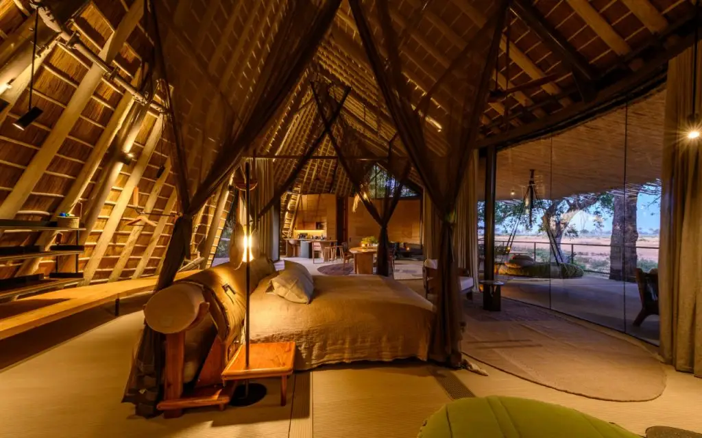Spacious thatched safari suite with luxurious four-poster bed looking out across a shaded balcony onto the Okavango Delta.