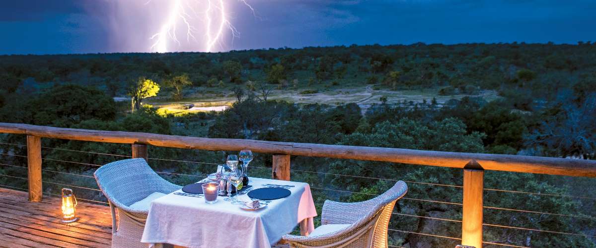 Lantern lit private dinner on wooden deck overlooking spot-lit watering hole with lightning bolt in the distance..