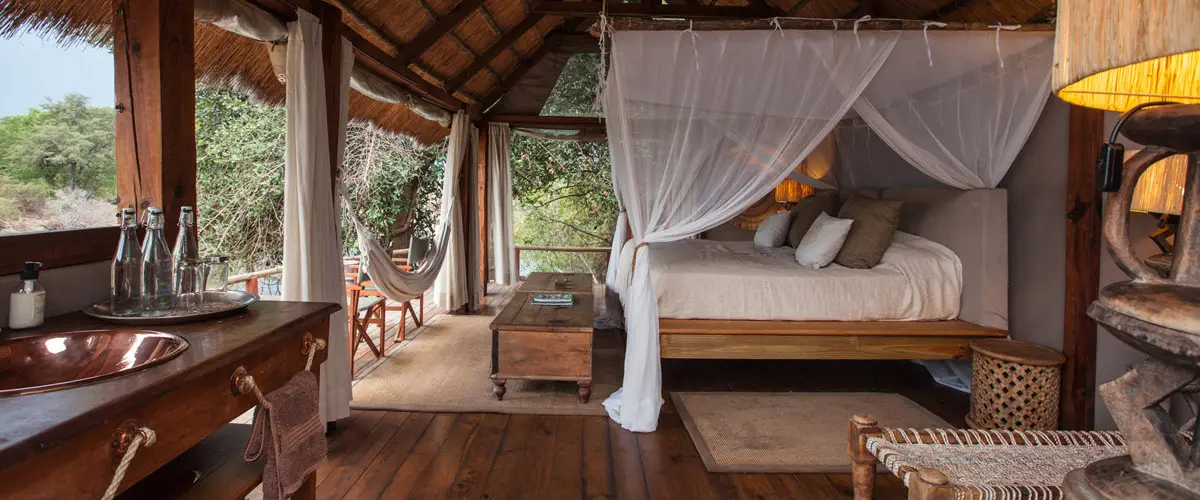 Open sided thatch and wood suite with four-poster bed leading onto balcony with hammock at Sindabezi Island Lodge