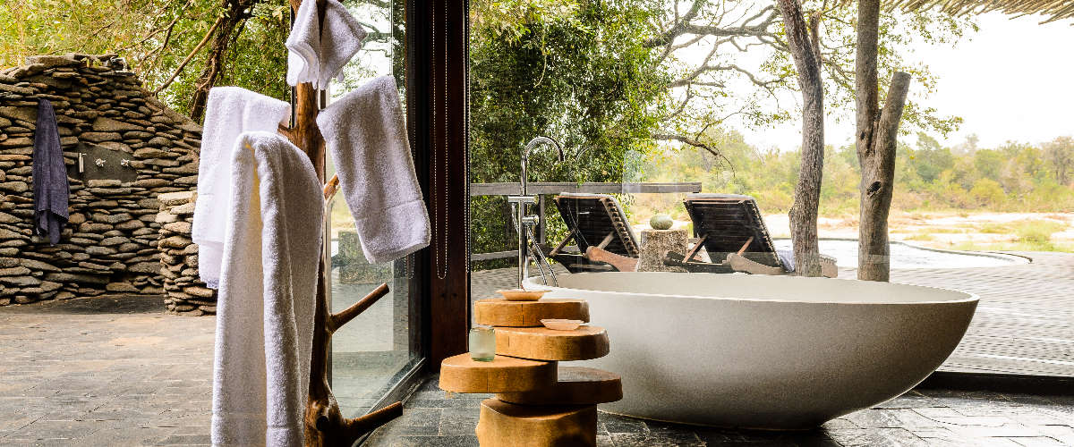 Bathroom with outdoor shower, freestanding tub and large windows framing the African bush.