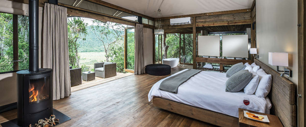 Luxury safari tent interior with modern bedroom, fireplace, open bathtub and outdoor balcony with distant views.